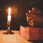 Close up man's hand is praying in the Church with lit candle, religion and faith concept